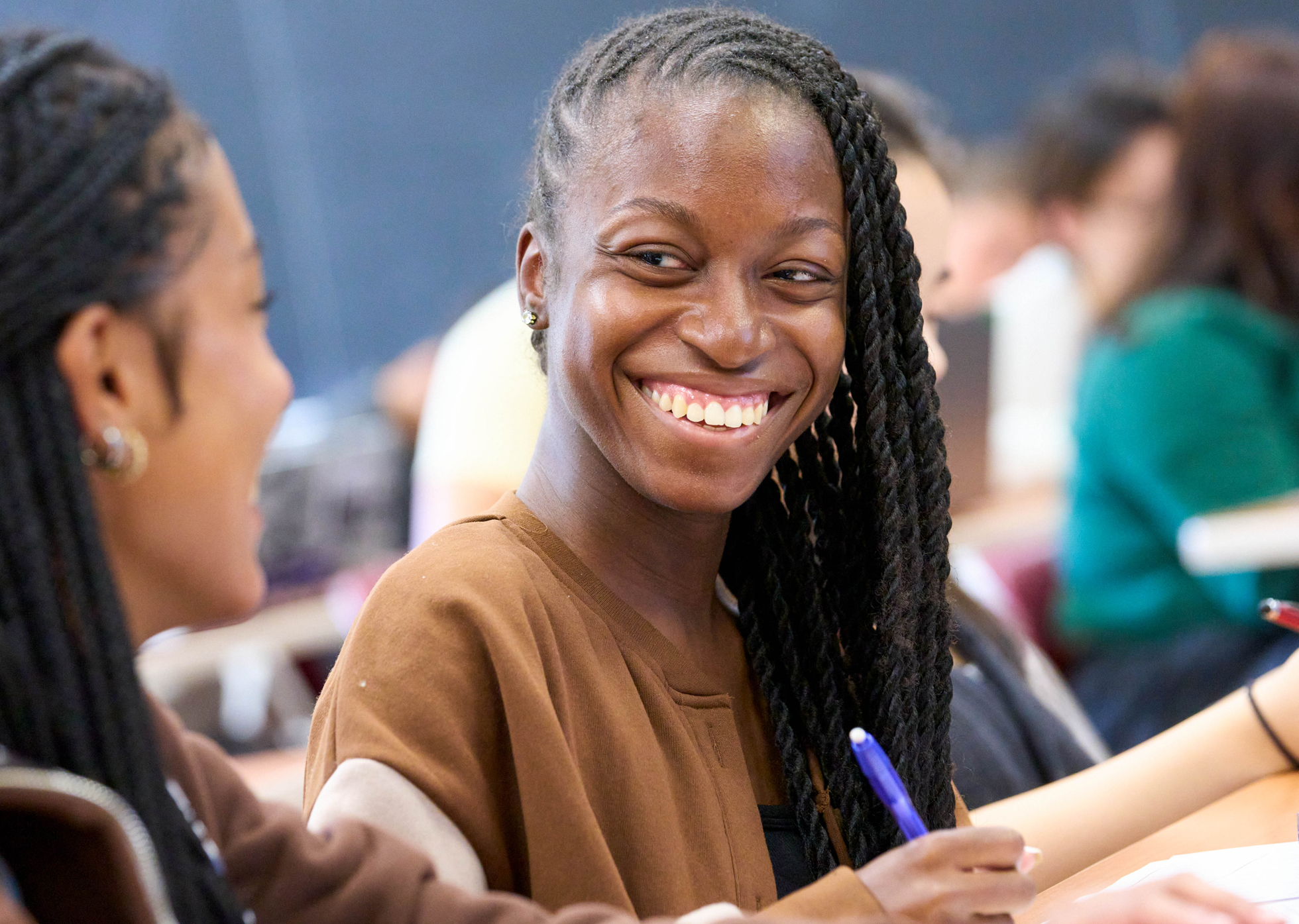 A young woman with braided hair smiles in a classroom setting, holding a blue pen