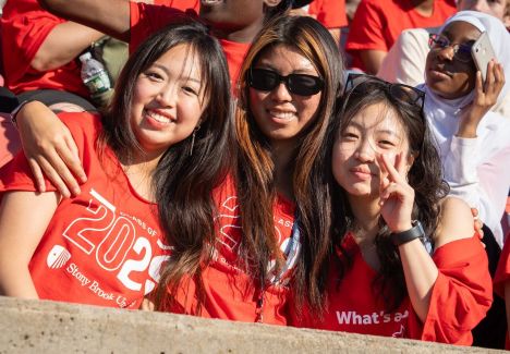 Three young women in red shirts smiling together, one wearing sunglasses and another showing a peace sign, at a festive event.