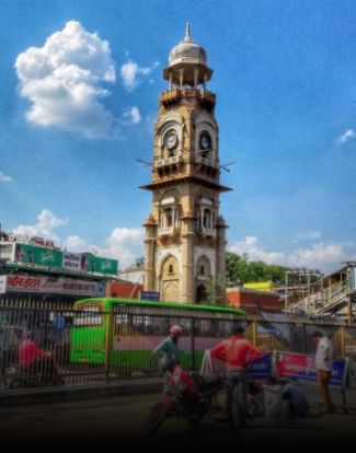 A tall, ornate clock tower stands against a blue sky with clouds, surrounded by people and commercial buildings.