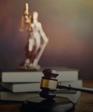 A gavel with a gold band rests on a circular base in the foreground, with a blurred statue of Lady Justice in the background, holding scales and a sword. Stacked books are visible behind the gavel.