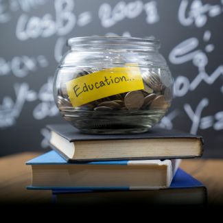 A glass jar filled with coins sits on top of three stacked blue books, with a yellow sticky note that reads "Education..." The background features a blackboard with white mathematical equations.