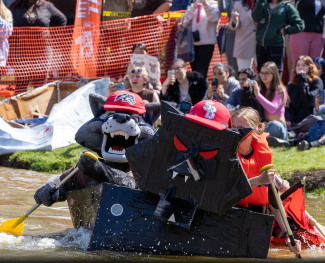 Stony Brook University mascot, Wolfie and participants in paddle a black cardboard boat with a fierce face during a lively boat race. A crowd watches from the shore.