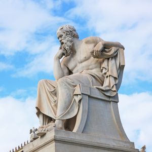 Marble statue of a seated philosopher in a thoughtful pose, resting his chin on one hand with a scroll in the other, set against a partly cloudy blue sky