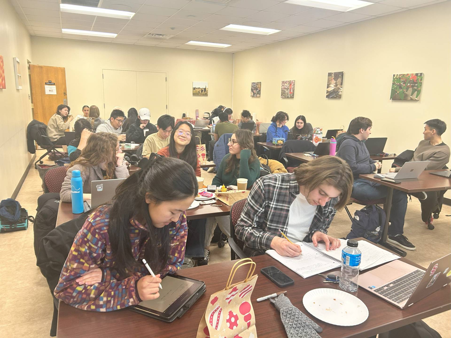 Students in a classroom setting working and studying at tables with laptops and tablets.