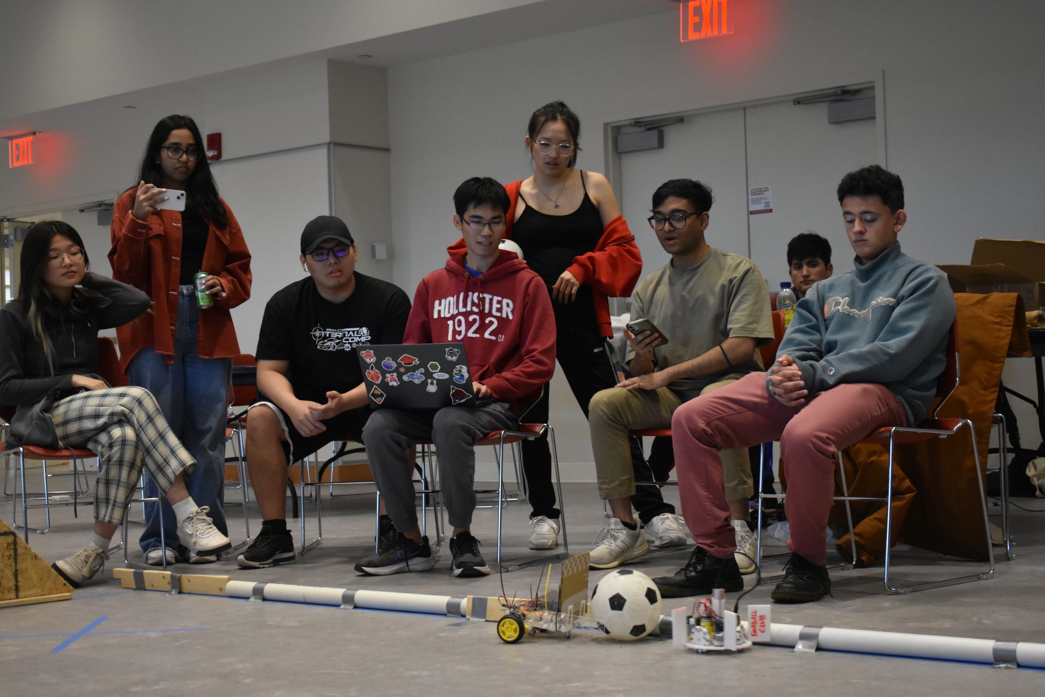 A group of students sitting down moving a remote controlled robot with a soccer ball.