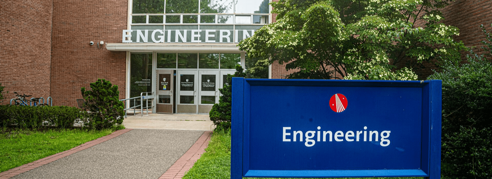 Entrance of an engineering building with a path leading to glass doors. A blue sign with "Engineering" stands in the foreground, surrounded by greenery.