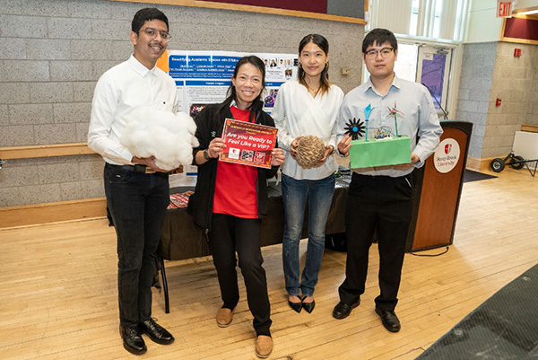A professor and three students are smiling in front of their table which demonstrates their project and they are holding items 