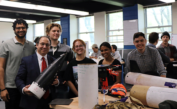 A group of six people stand in a classroom, smiling at the camera. One person holds a rocket component.
