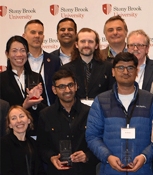 A diverse group of people smiling and holding awards in front of a Stony Brook University backdrop.