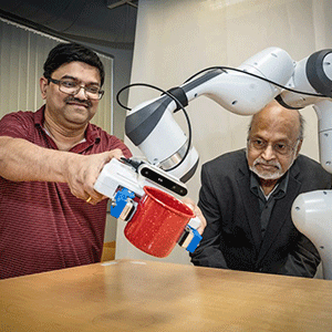 Two men interact with a robotic arm holding a red mug. One man observes intently; the other assists.