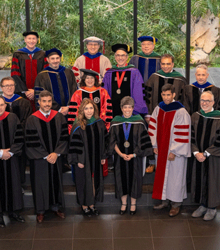 A group of 15 people in academic regalia, including caps and gowns, pose indoors against a backdrop of lush greenery, conveying a celebratory tone.