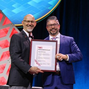 Two men smiling while holding a framed award certificate on stage, with a geometric backdrop.