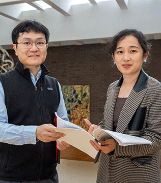 A man and woman stand indoors, engaging with open notebooks.