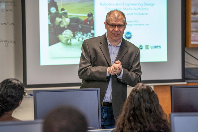 Professor Jon Longtin in front of his classroom with a slide behind him of a mechanical engineer at work