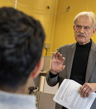 A man with a gray blazer gestures while holding documents, engaging with a person in the foreground. The room has yellow walls, conveying a serious tone.