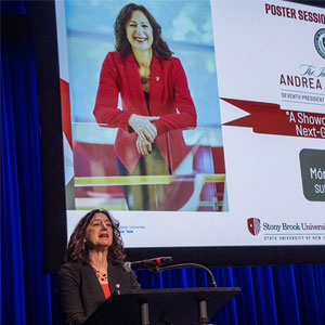 A woman stands at a podium, speaking in front of a large screen displaying a poster session for Stony Brook University.