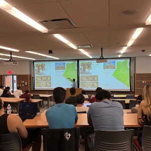 A classroom with a teacher presenting at the front to students seated at desks, with two large screens displaying information.