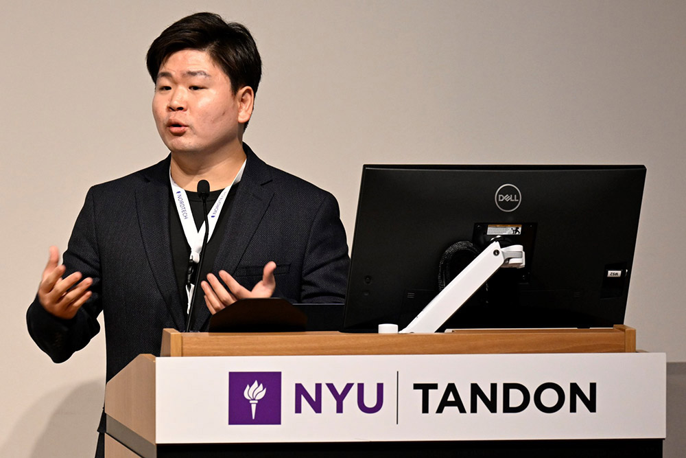 A man in a black suit is speaking at a podium with a computer to his right.