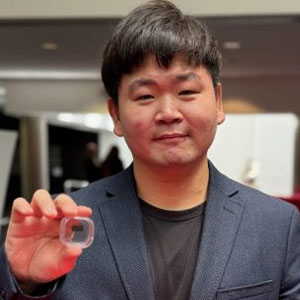 Man in a dark blazer holding a small transparent object toward the camera.
