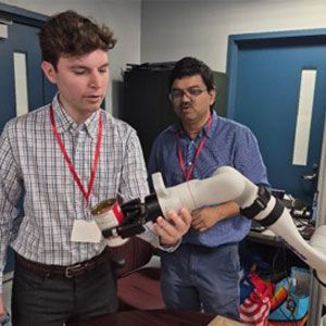 Two men examine and hold a white prosthetic limb in an indoor clinical or technical setting.