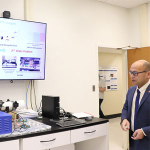A man in a suit stands in an electronics laboratory beside a workbench and a display showing a slide.