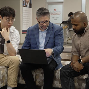 Three people sitting on a bench, looking at a laptop together.