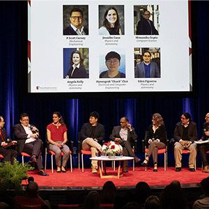 A diverse group of eight people sits on stage, engaged in a panel discussion. A presentation with six headshots and names is displayed on a screen above.