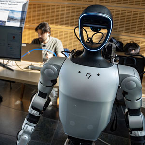 A humanoid robot with a sleek design in the foreground, softly lit, stands in a tech lab. A person working on a computer is visible in the background.