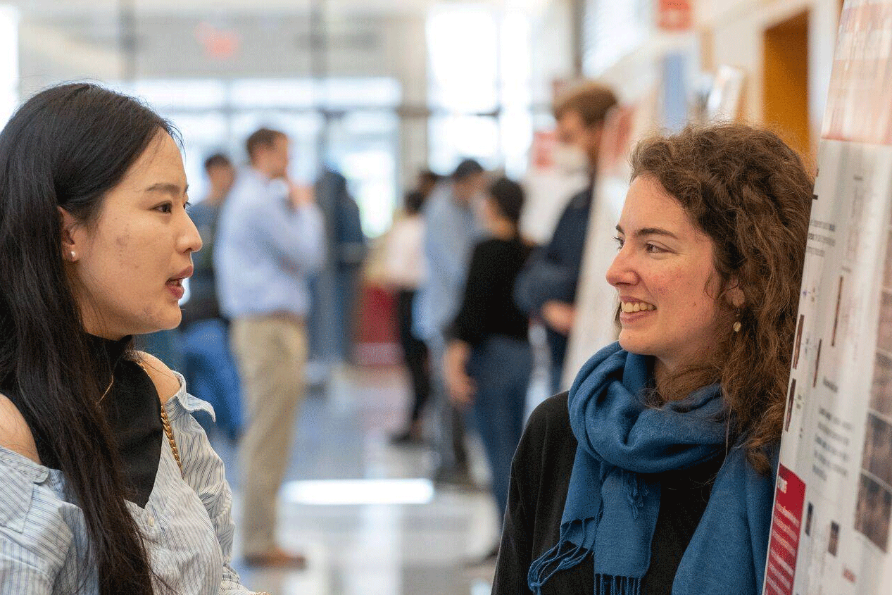Two women are conversing at a poster presentation in a bright hallway. One holds a drink, and both are smiling.