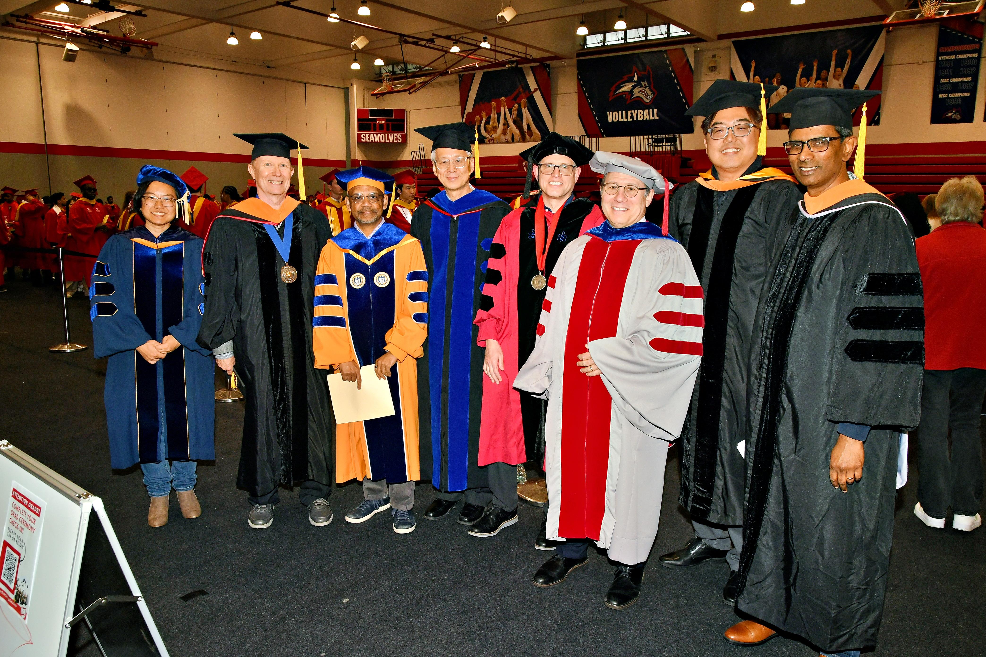 A group of eight academic professionals in colorful graduation robes and caps stand smiling in a gymnasium
