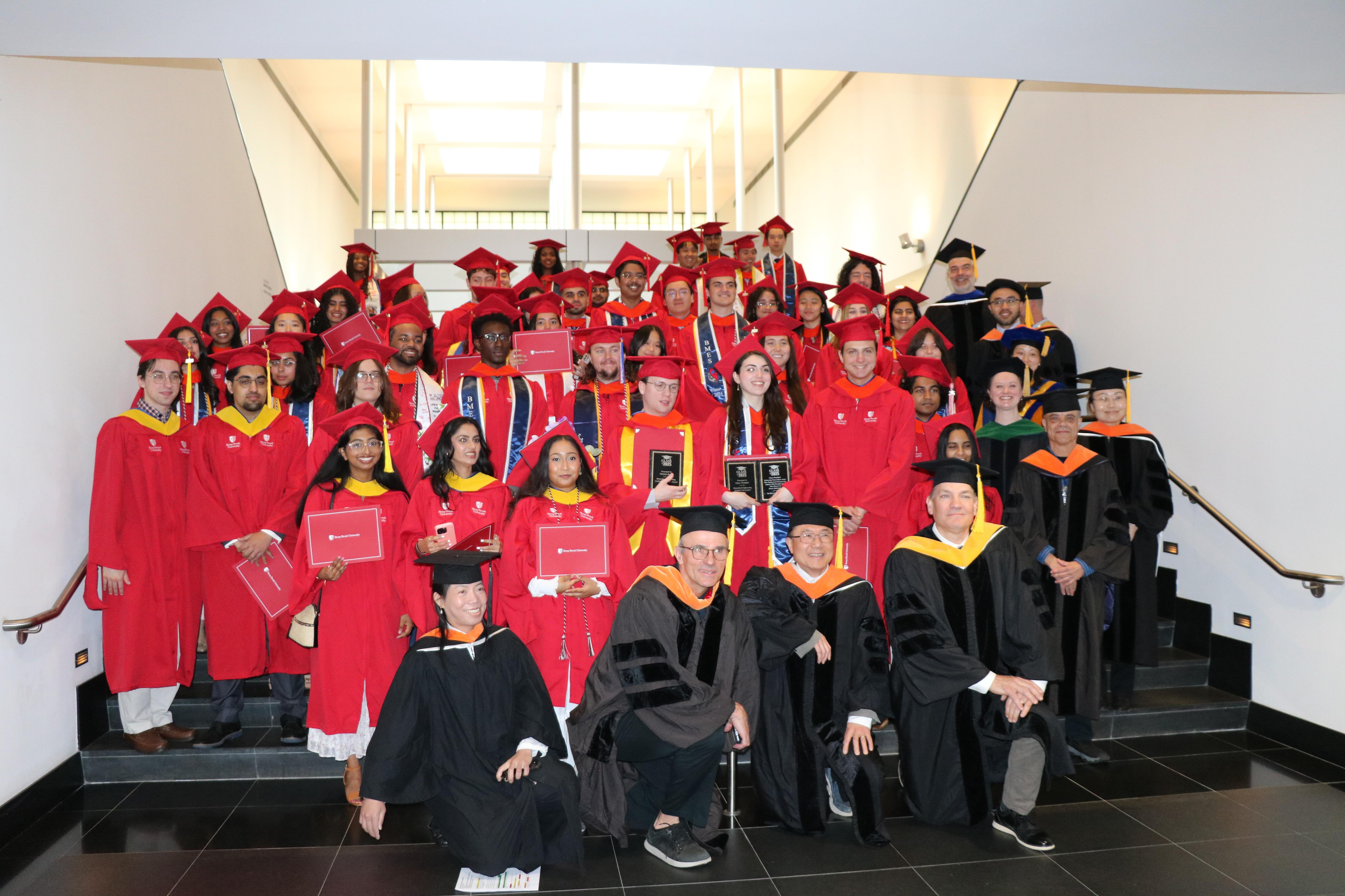 Graduates in red gowns and caps stand on stairs, holding diplomas and awards. Faculty in black robes surround them, celebrating with smiles.
