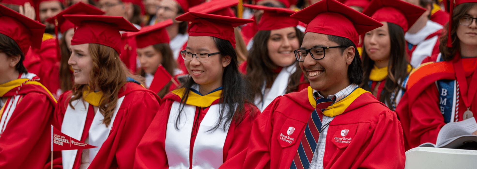 Two students sitting in the crowd smiling at Commencement in red cap and gowns.