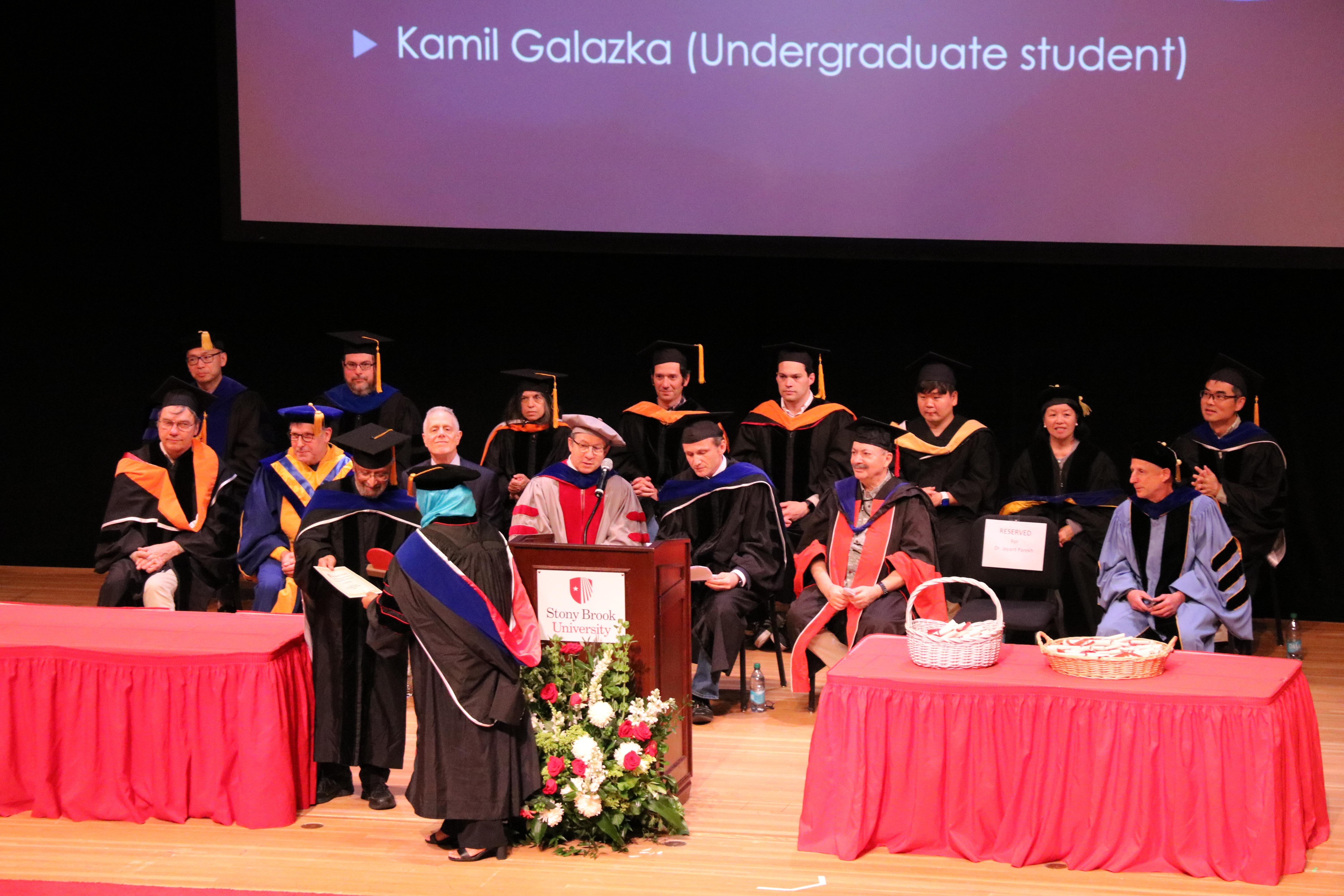 A graduation ceremony with faculty on stage in academic regalia. A student is recognized at a podium with a projected name on a screen. 