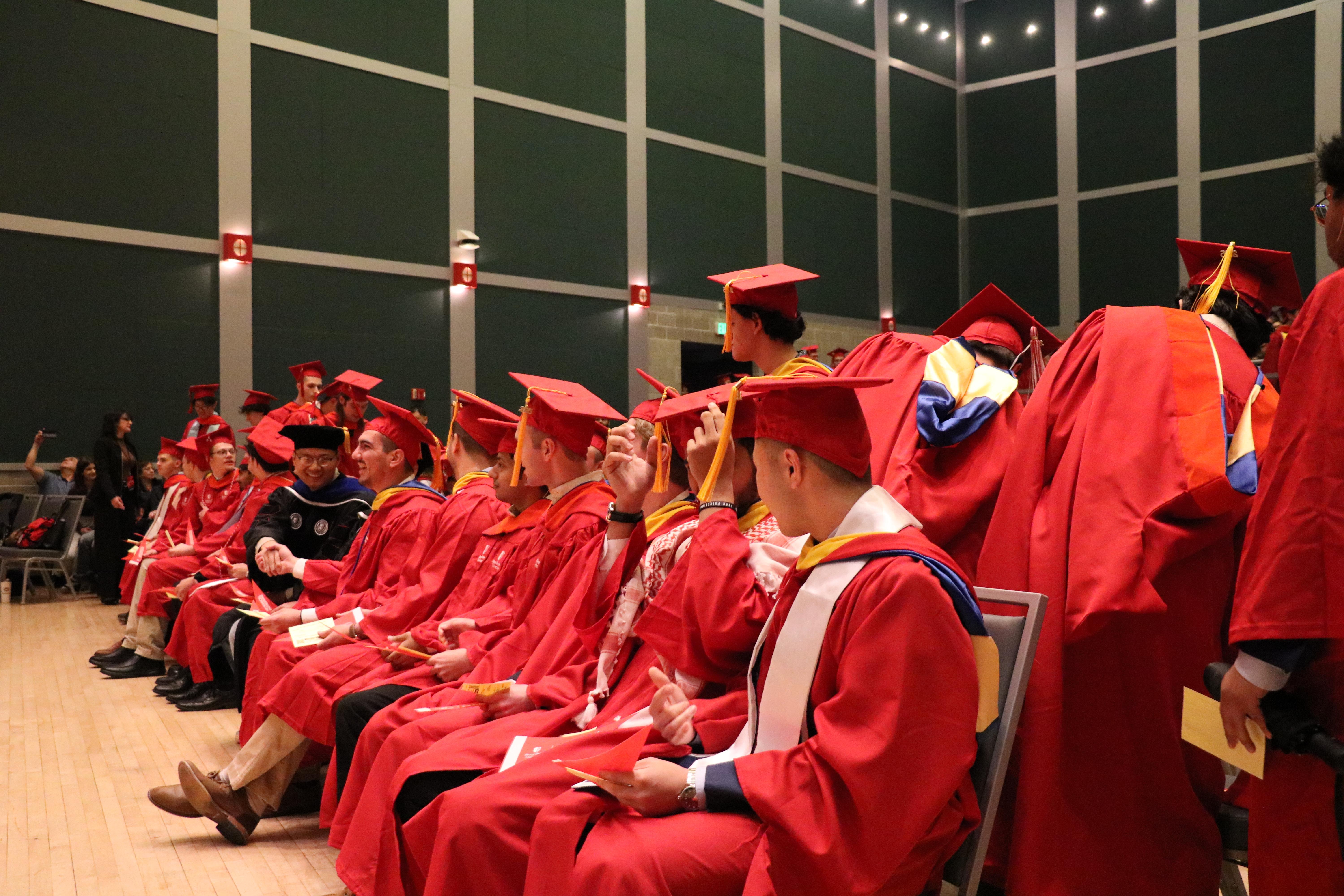 Graduates in red caps and gowns sit in rows, some adjusting tassels.