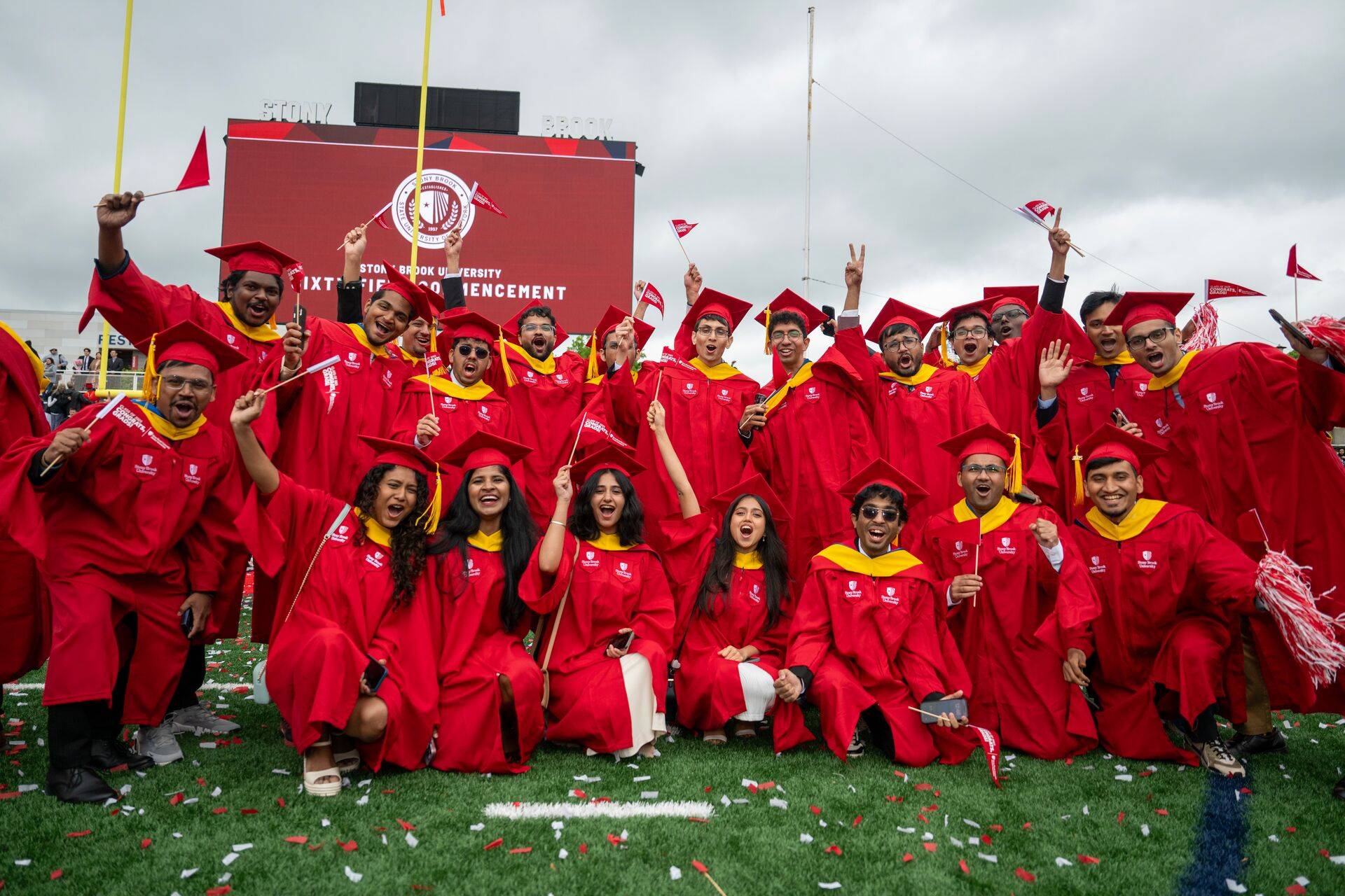 Graduates in red gowns and caps celebrate on a field, waving flags and cheering. A large screen in the background displays "Commencement."
