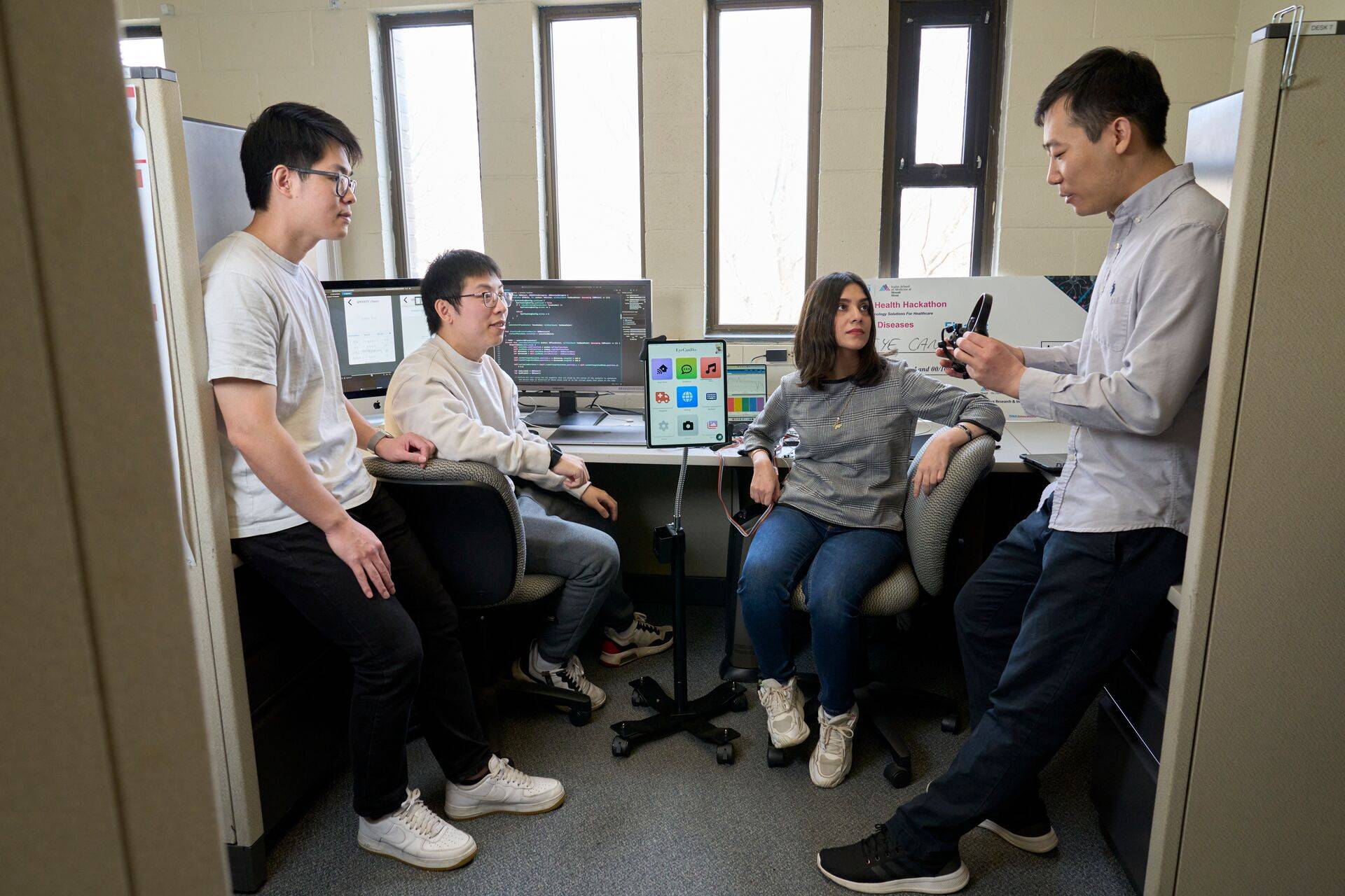 A group of four people in an office, engaged in discussion. Two are seated at desks with computers, displaying code and apps.
