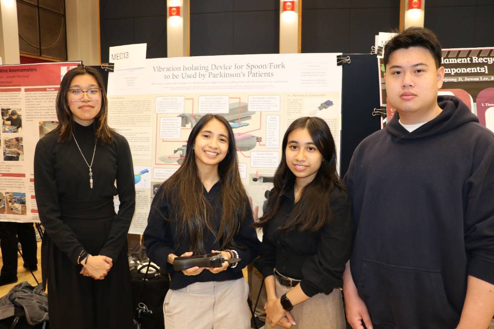 Four students stand proudly in front of their science project display on a vibration isolating device for Parkinson's patients, conveying teamwork and innovation.