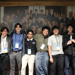 A group of six smiling men, casually dressed with conference badges, pose in front of a large historical painting depicting a formal gathering of diplomats.