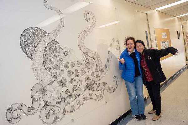 Two people smile and pose next to a detailed black-and-white octopus drawing on a wall.