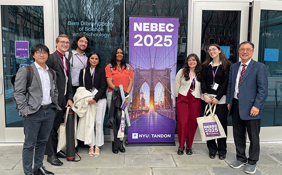 A group of eight people stand smiling in front of a building, next to a banner reading "NEBEC 2025 NYU Tandon" with a Brooklyn Bridge image.