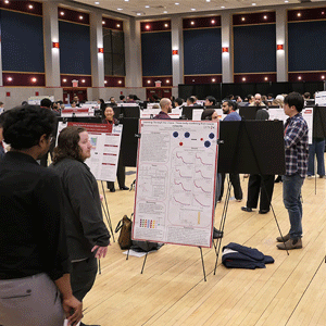 People attend a busy poster session in a large hall, engaged in discussions around various displayed research posters. The atmosphere is collaborative.