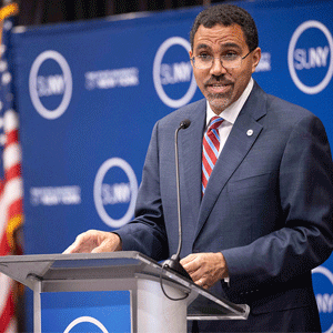 A man in a suit and red-striped tie speaks at a podium with a microphone. Behind him is a blue SUNY backdrop and a U.S. flag.