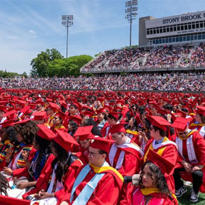 Students in red cap and gowns sitting on a field during Commencement.
