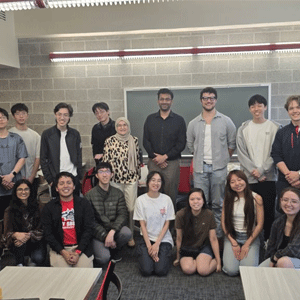 A diverse group of 16 people, some standing and some kneeling, smiling in a classroom with a brick wall and chalkboard, conveying a cheerful atmosphere.