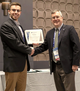 Two men in suits shake hands, smiling, as one holds an award certificate. They stand in a professional setting, with a patterned wall behind them.