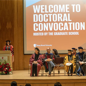 People on stage in graduation cap and gowns with screen behind stating "Welcome to Doctoral Convocation."