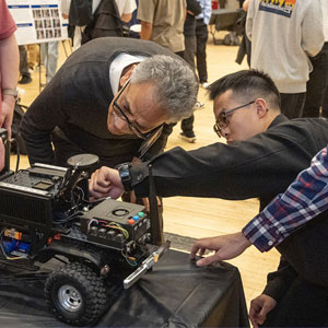 Two people closely examine a small, wheeled robotic vehicle on a table. They appear focused and engaged, with others in the background at an event.