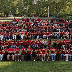 A large group of people in red shirts seated in rows on a grassy area, holding hands, symbolizing unity.