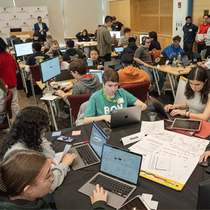 A diverse group of young people working at laptops around tables in a busy, collaborative environment, suggesting teamwork and concentration.
