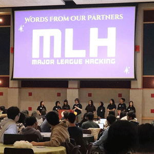 Projection of "words from our partners MLH, Major League Hacking" on screen above a group of students.